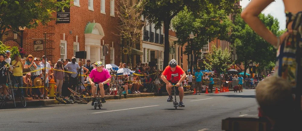 Two children riding scooters in a street race during a community event, with spectators lined along the sidewalk and trees providing shade.