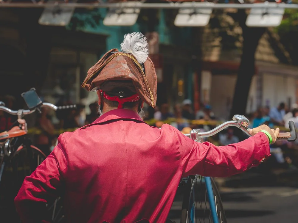 Back view of a person wearing a historical costume with a tricorn hat and red jacket, holding a bicycle in a busy outdoor setting.