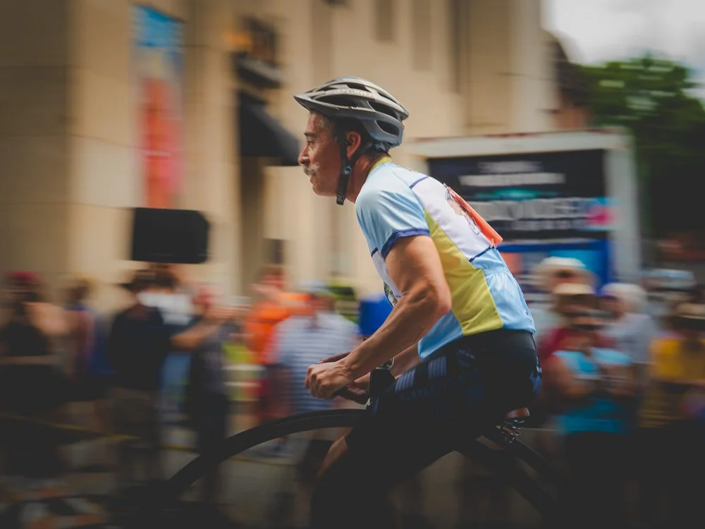 Older man riding a bicycle during a public event or race, wearing a helmet and colorful athletic shirt, with a crowd of spectators blurred in the background.