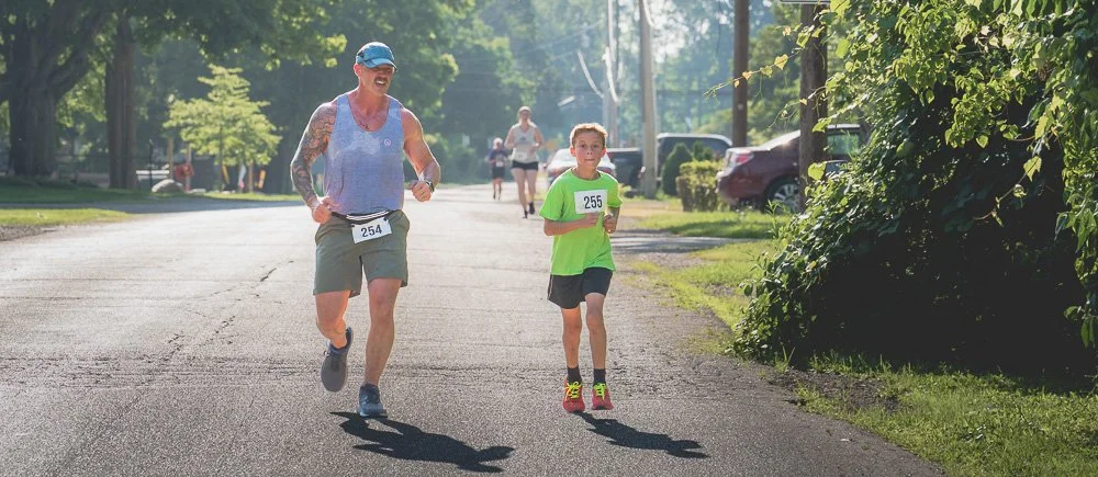 A man and a boy running in a road race on a sunny day, both wearing race bibs with numbers 254 and 255, surrounded by trees and parked cars.