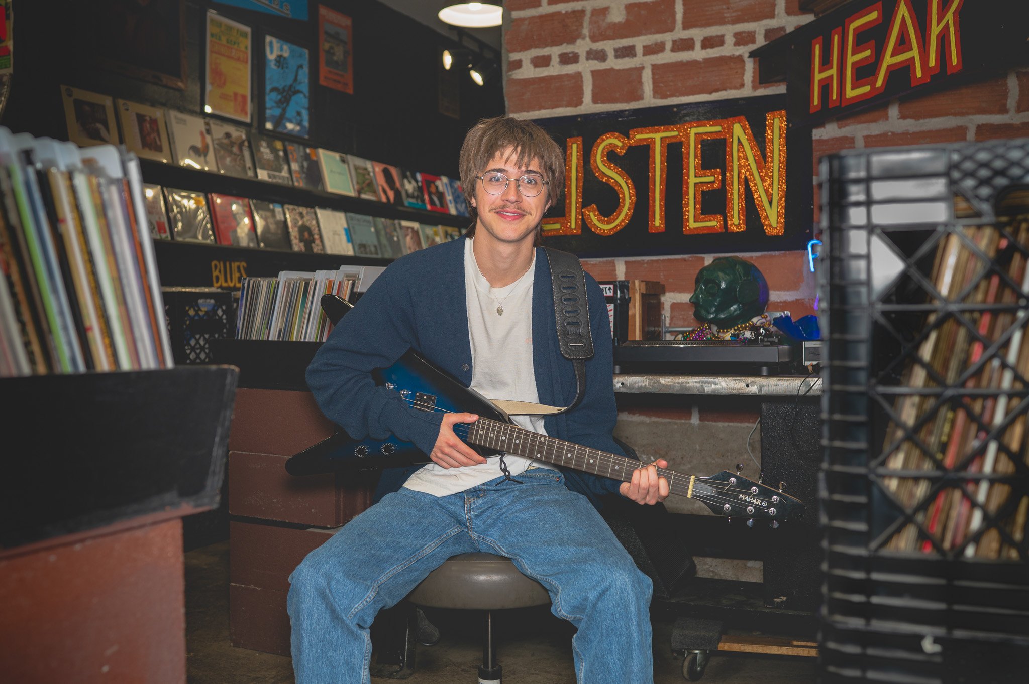 A young man with glasses and a mustache sitting on a chair in a record store, holding an electric guitar. Behind him, a neon sign reads 'LISTEN' and 'HEAR' on a brick wall, with shelves of vinyl records and books around.