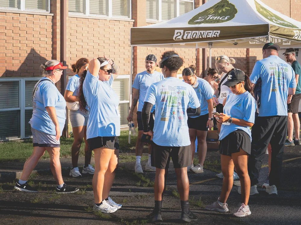 Group of people gathering under a tent, wearing matching light blue T-shirts, outside a brick building on a sunny day.