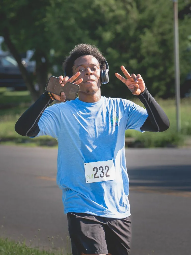 Young man running in a race, wearing a blue t-shirt, black sleeves, and a race bib numbered 232, while listening to music with headphones and making peace signs with both hands.