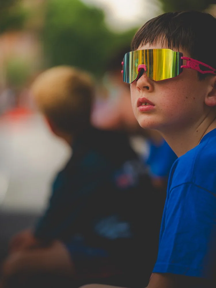 Close-up of a young boy with dark hair wearing reflective, multicolored goggles and a blue shirt, sitting outdoors with blurred background and other children.