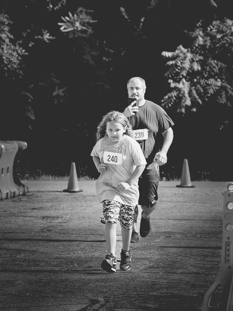 A young girl and an older man running together in a race, wearing numbered bibs, on an outdoor track with cones and trees in the background.