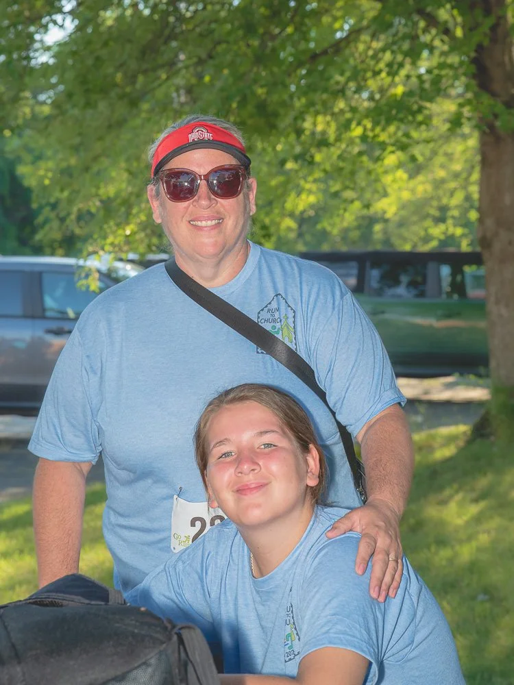 A woman wearing sunglasses, a red Ohio State headband, and a blue t-shirt standing outdoors with her arm around a young girl in a blue t-shirt, both smiling. Background includes green trees and parked cars.