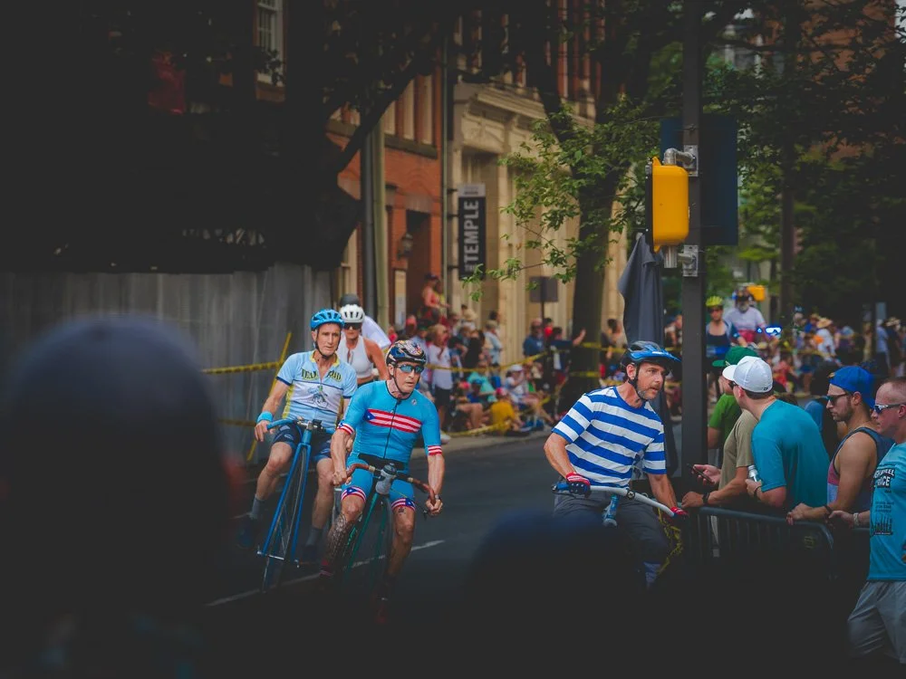 Cyclists riding bikes on a city street during a race, with spectators watching behind barriers and trees lining the street.