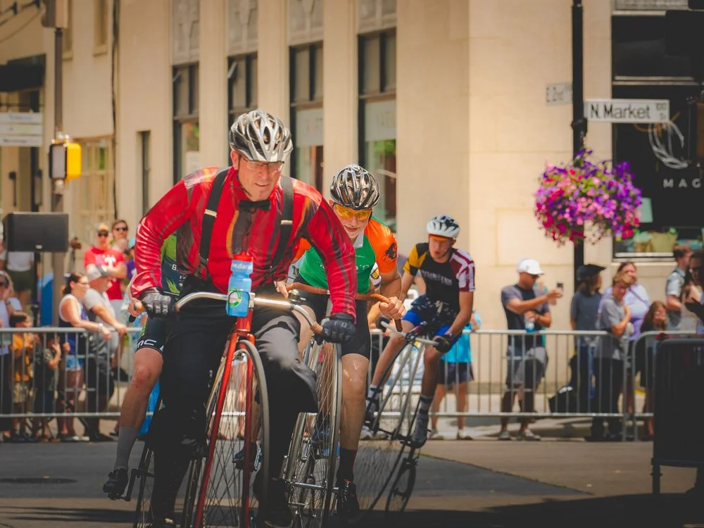 Four men, wearing helmets and casual athletic clothes, participating in a penny-farthing bicycle race on a city street with spectators behind barriers.
