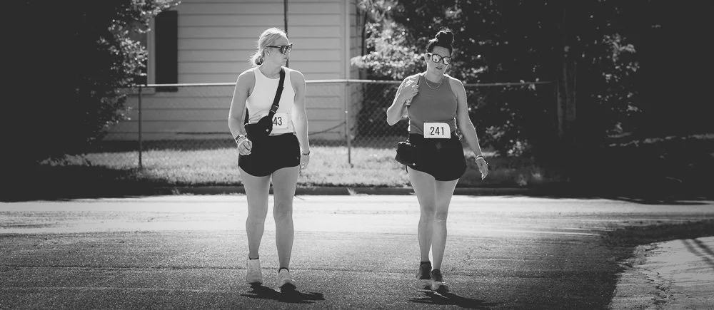 Two women in athletic wear and shorts walking on a city street, each with a race bib and a fanny pack, under sunlight.