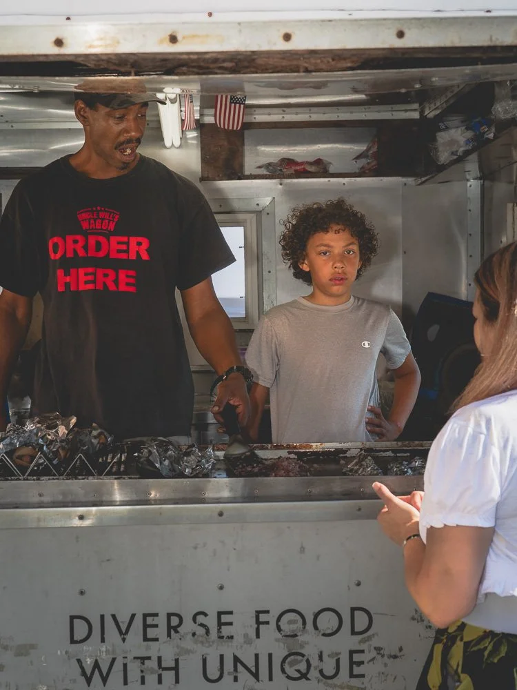 A man and a boy stand behind a food truck window, with a woman in front of them. The man is wearing a black shirt with red text that says "Order Here" and a brown hat. The boy has curly hair and is wearing a gray T-shirt. The woman is pointing at the