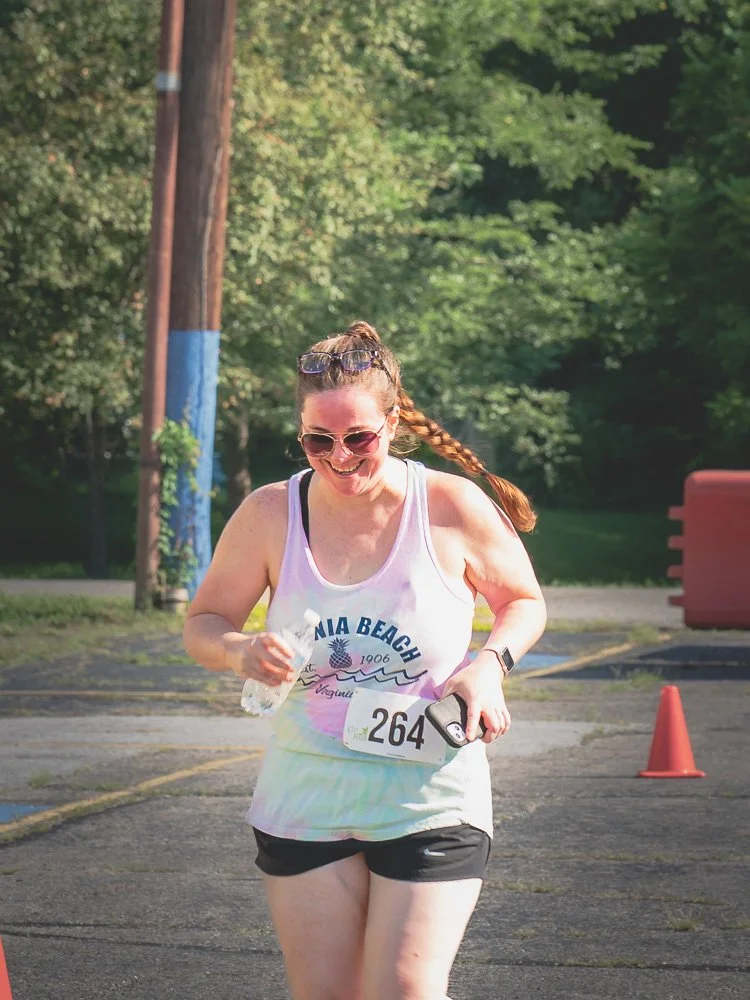 Female runner smiling and holding water bottle and phone during outdoor race, wearing sunglasses, a racerback tank top with 'Virginia Beach' on it, black shorts, and race bib number 264, with trees and orange cones in the background.