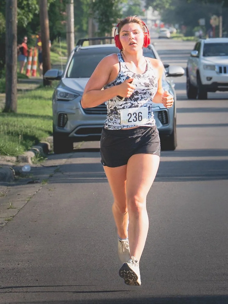 Young woman running in a race, wearing a tank top, shorts, and large red headphones, with race bib number 236, on a city street.