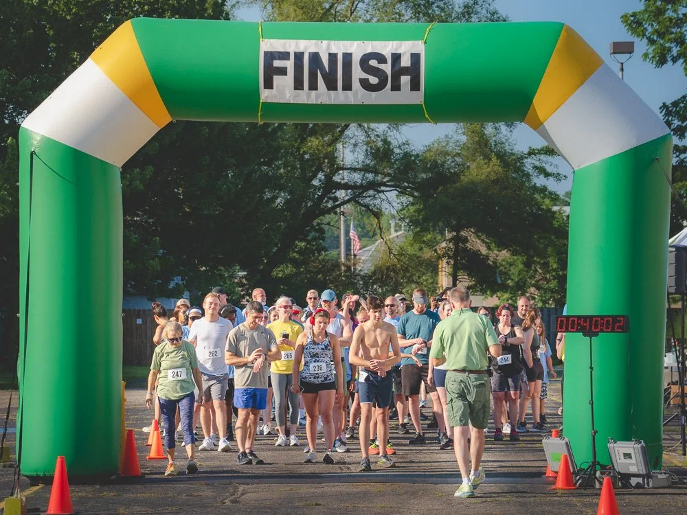 Group of people at the starting line of a race under a green and yellow inflatable finish arch, with some runners wearing bib numbers and a digital timer displaying 4 seconds.