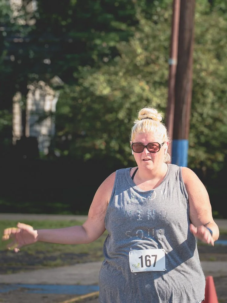 Woman with blonde hair in a bun, wearing sunglasses and a gray tank top, participating in a race with bib number 167, outdoors on a sunny day.