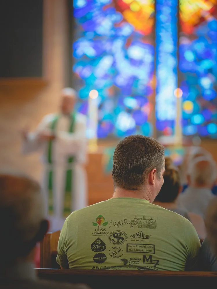 Man sitting in church pew with choir or speaker on stage and colorful stained glass window in background.