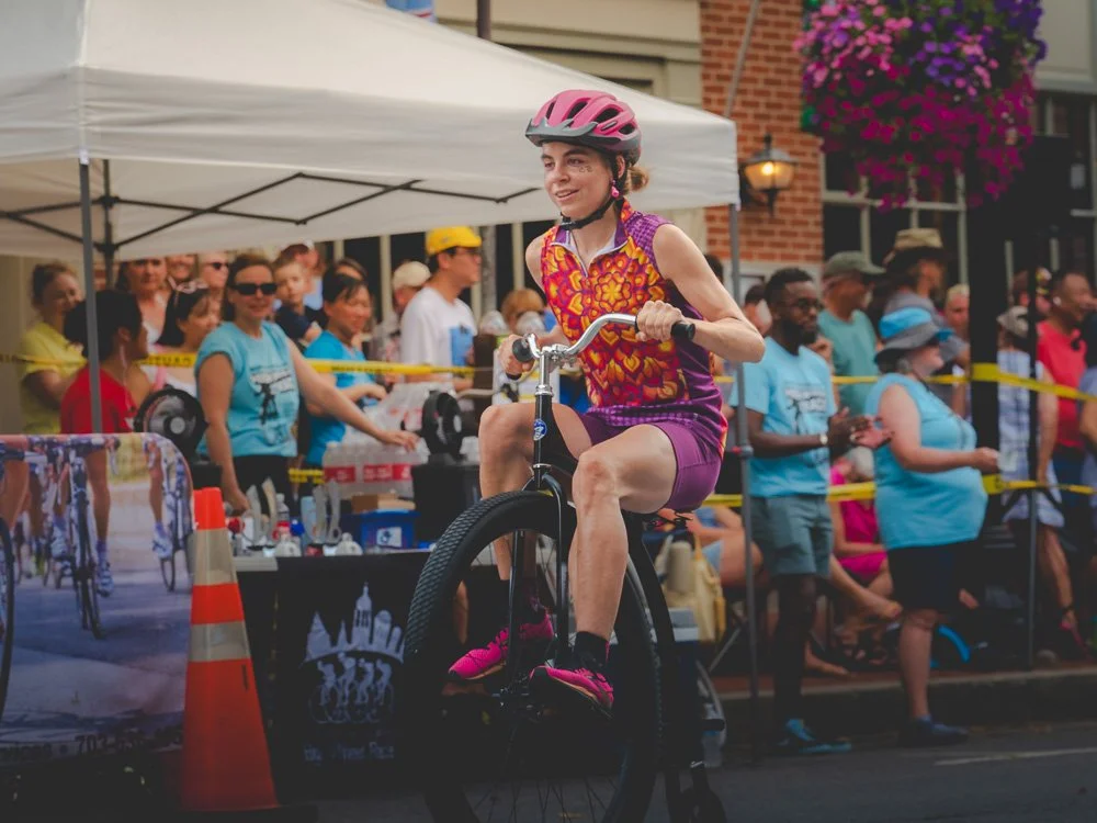 Woman riding a bicycle at a race event with onlookers behind a yellow safety line