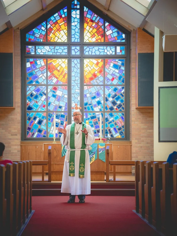 A priest in white robes with green stole standing at the altar of a church, with a large colorful stained glass window behind him and wooden pews on either side.