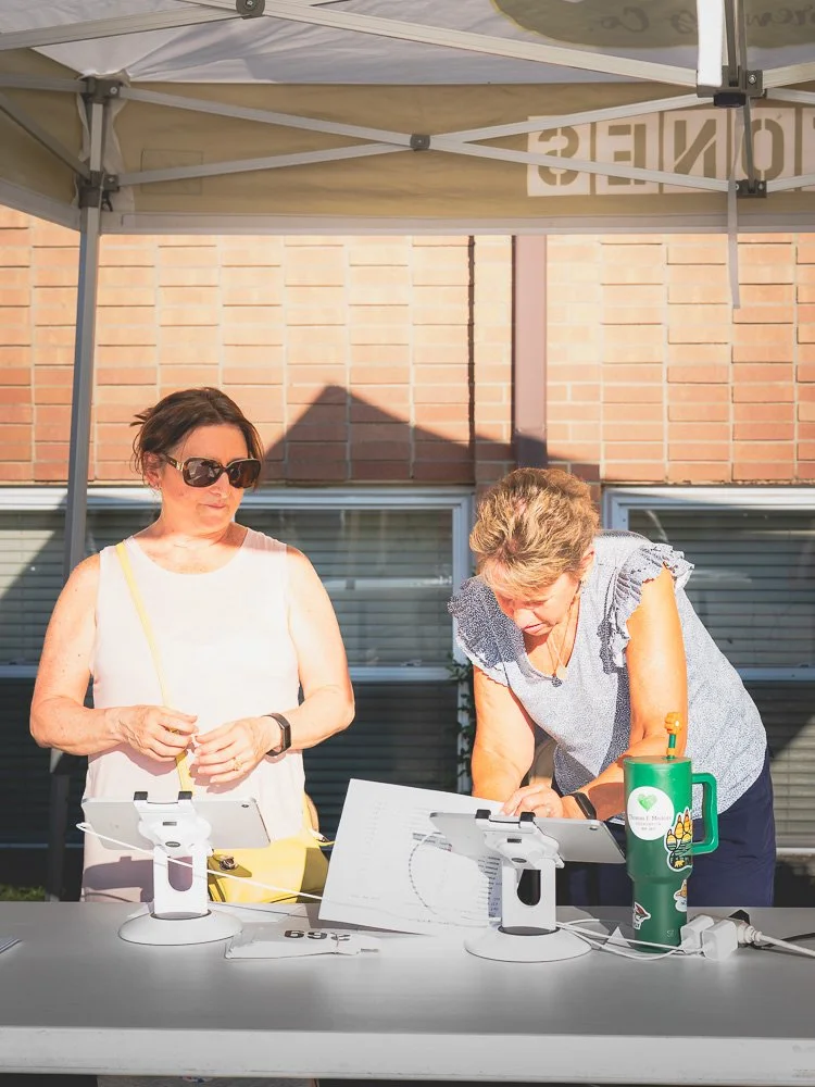 Two women standing at an outdoor table under a canopy, looking at tablets. One woman is wearing sunglasses and a white sleeveless top, the other woman is leaning over, looking at her tablet. There is a green water bottle on the table.