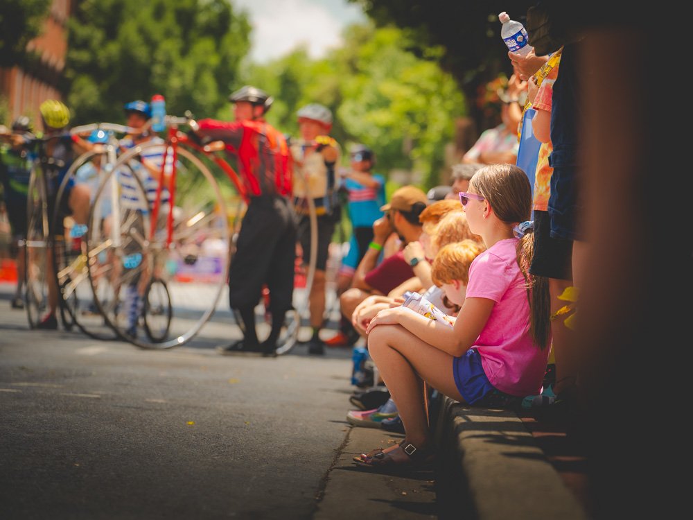 Children sitting on pavement watching a bicycle race, some holding water bottles, outdoors on a sunny day.