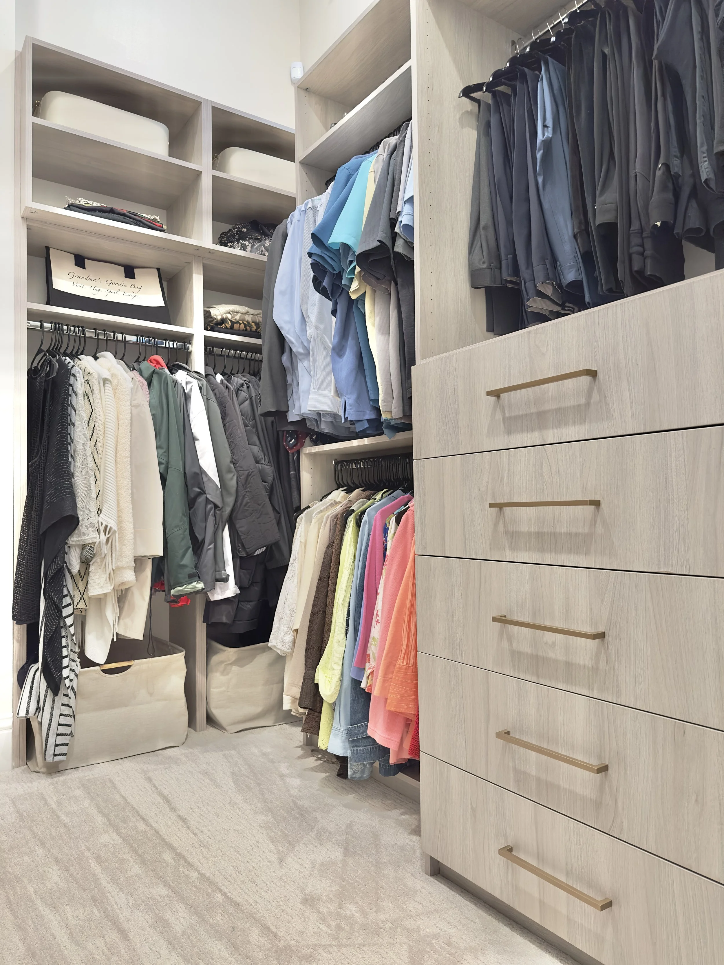 A walk-in closet with shelves, hanging clothes, and drawers. The closet contains various clothing items including jackets, shirts, pants, and T-shirts, organized by type and color. There are storage baskets on the floor and shelves on the wall.