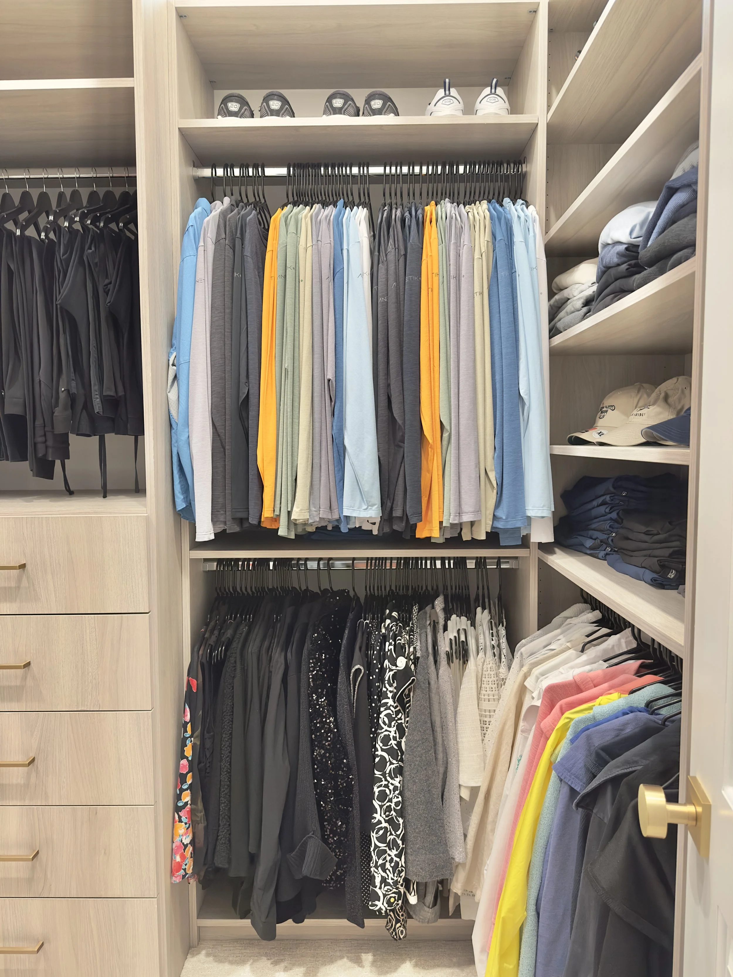 Closet with organized clothing, shoes, and accessories. The upper shelf has white and black shoes, hanging clothes are in various colors, and the bottom shelf has more clothes in dark and patterned fabrics.