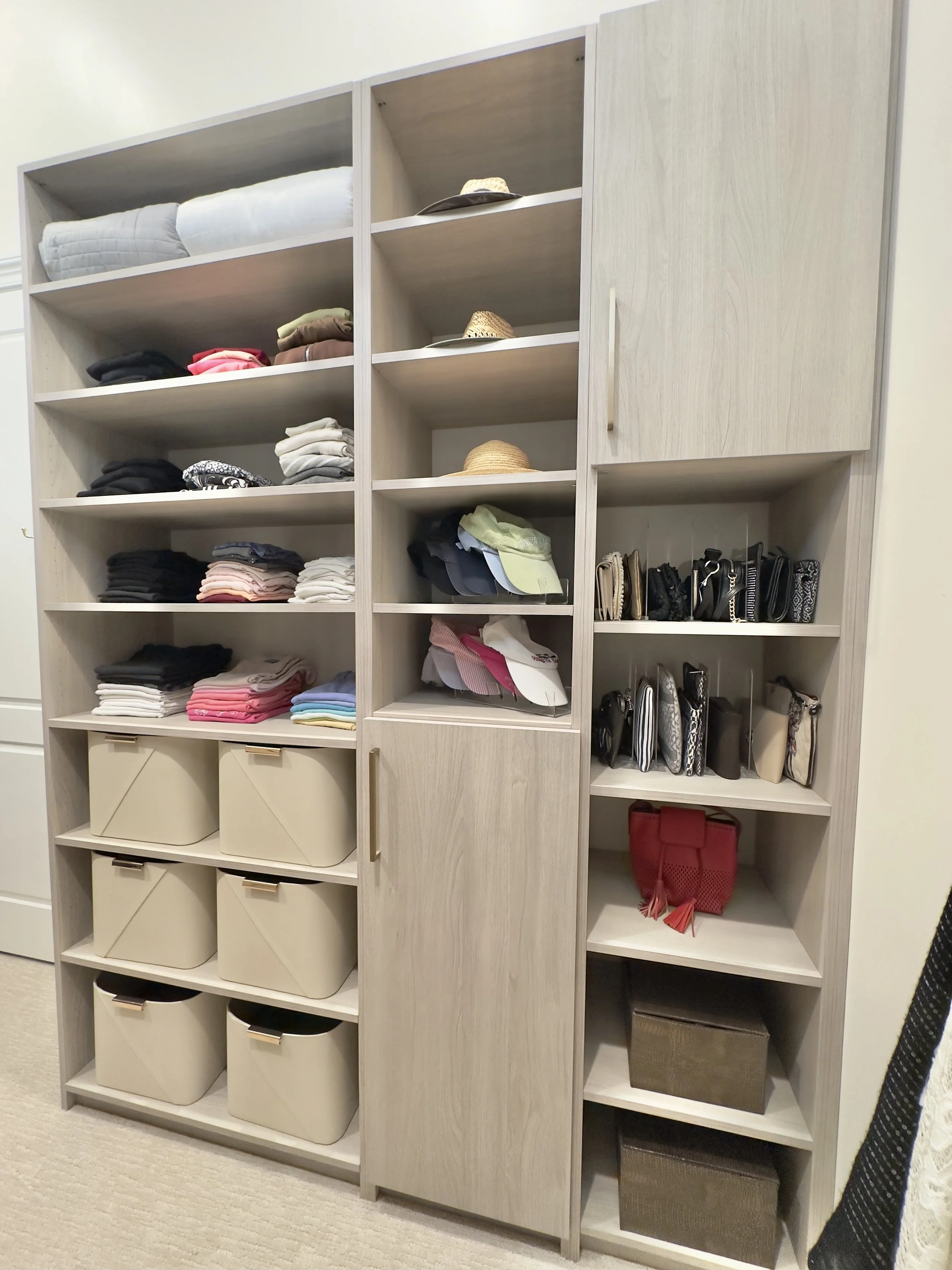 A tall wooden shelving unit filled with folded clothes, hats, handbags, and storage boxes in a neatly organized closet.