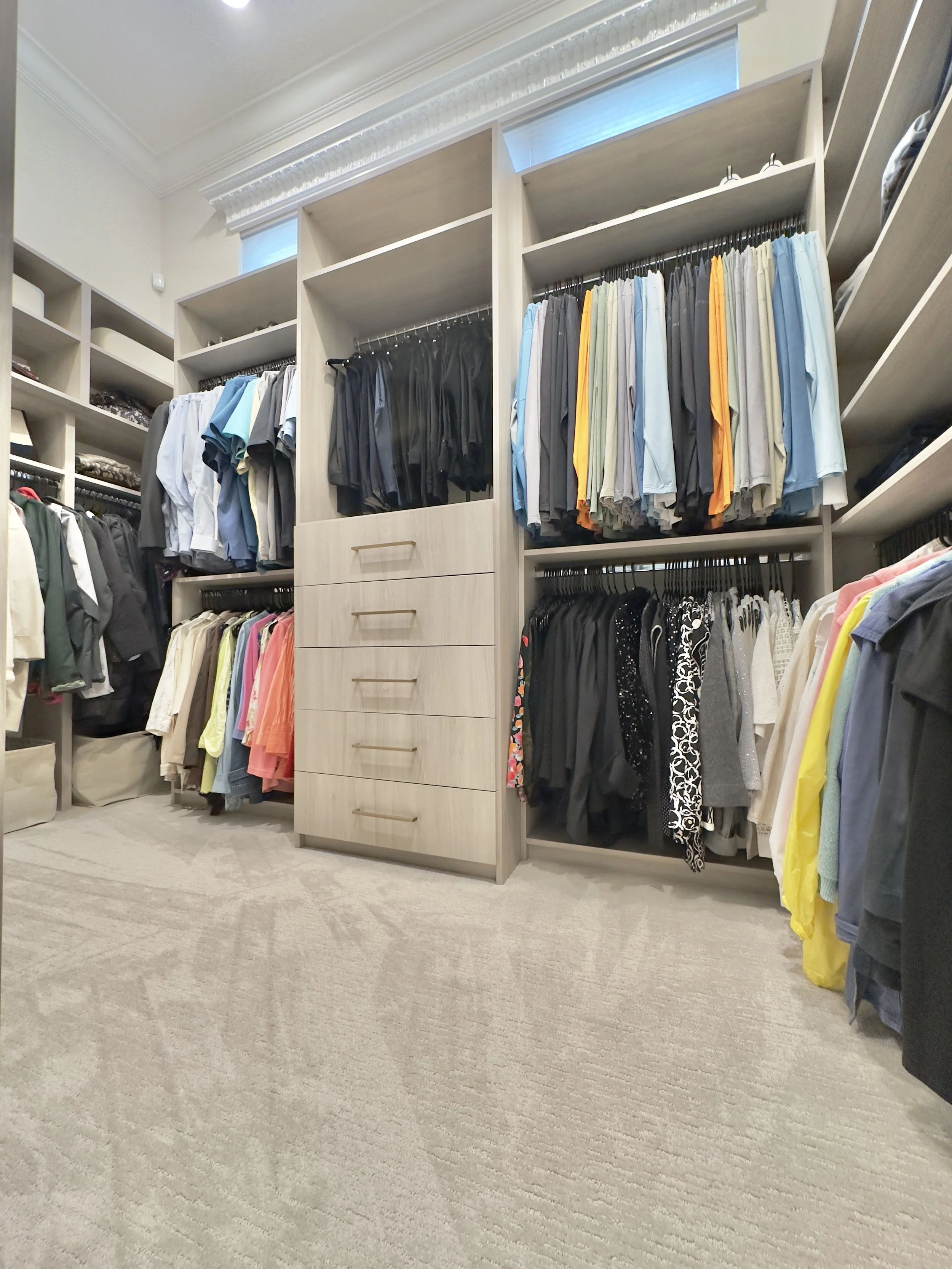 Spacious walk-in closet with organized clothing racks, drawers, and shelves filled with men's and women's clothes in various colors and styles.