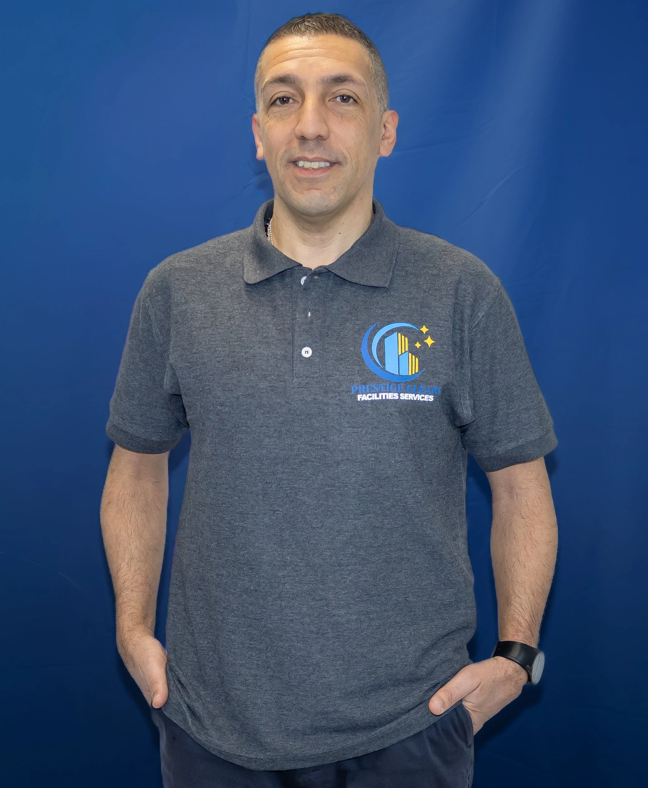 Man wearing a dark gray polo shirt with a logo that says 'Prestige Clean Facilities Services,' standing in front of a blue background with hands in pockets.