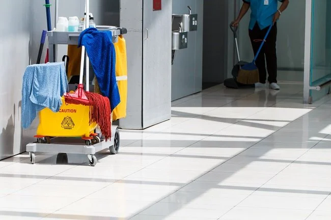 Cleaning cart with cleaning supplies and towels, and a janitor sweeping the floor in a building hallway.