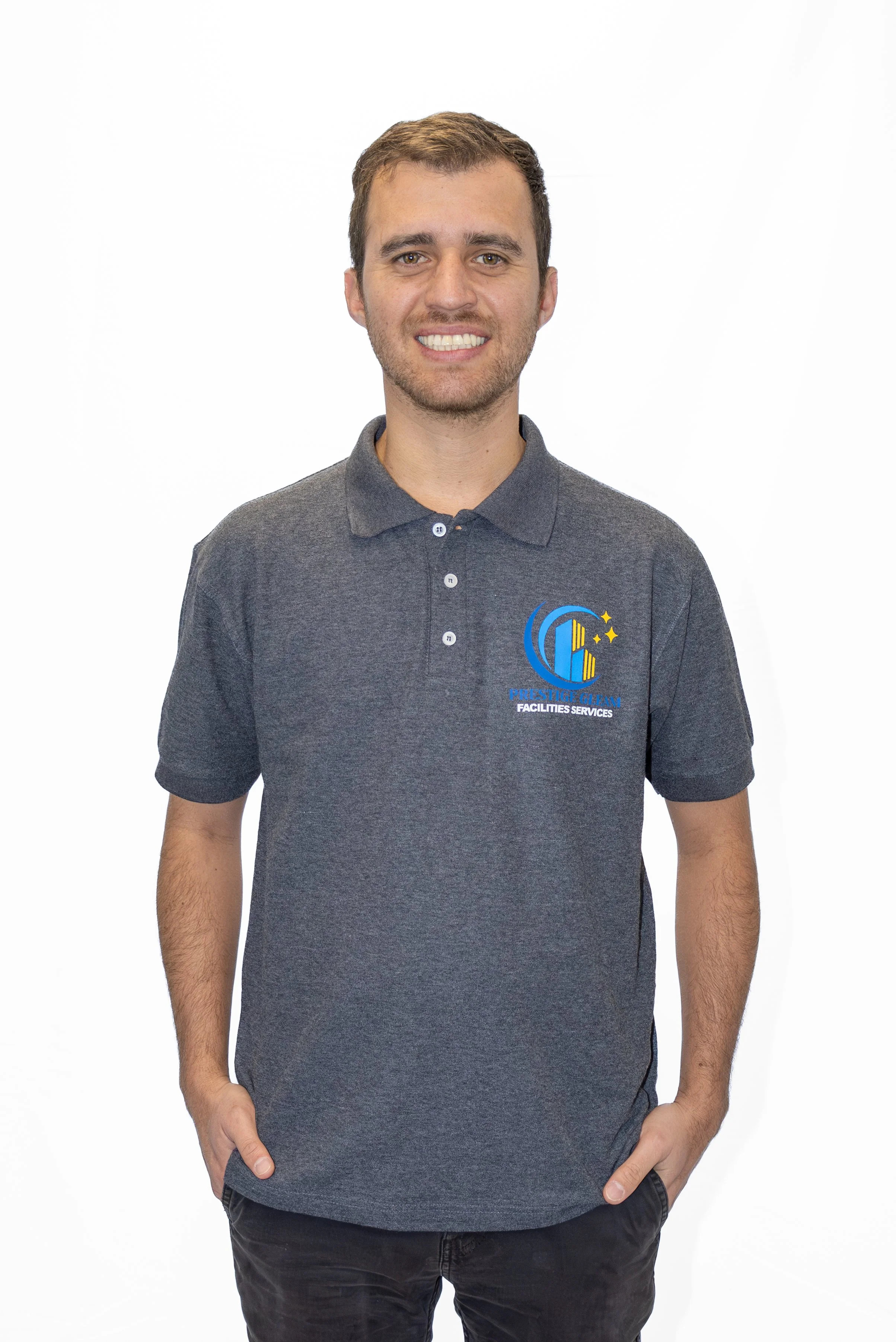 A young man with brown hair and a beard, wearing a dark gray polo shirt with a company logo on the chest, stands against a plain white background and smiles at the camera.