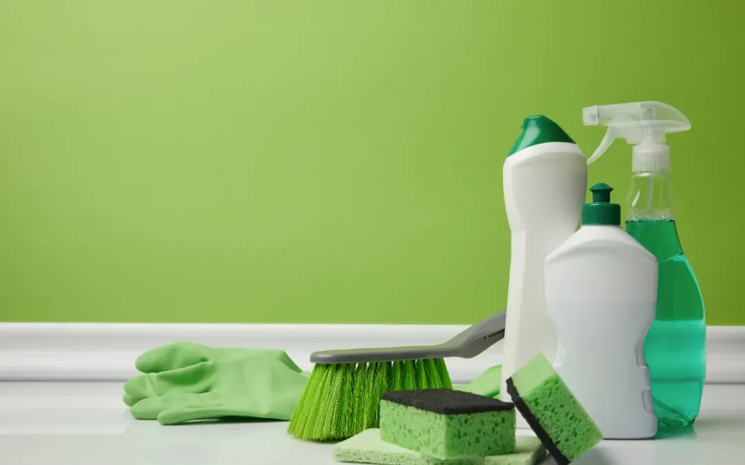 Cleaning supplies including spray bottles, sponges, a brush, and gloves against a green wall