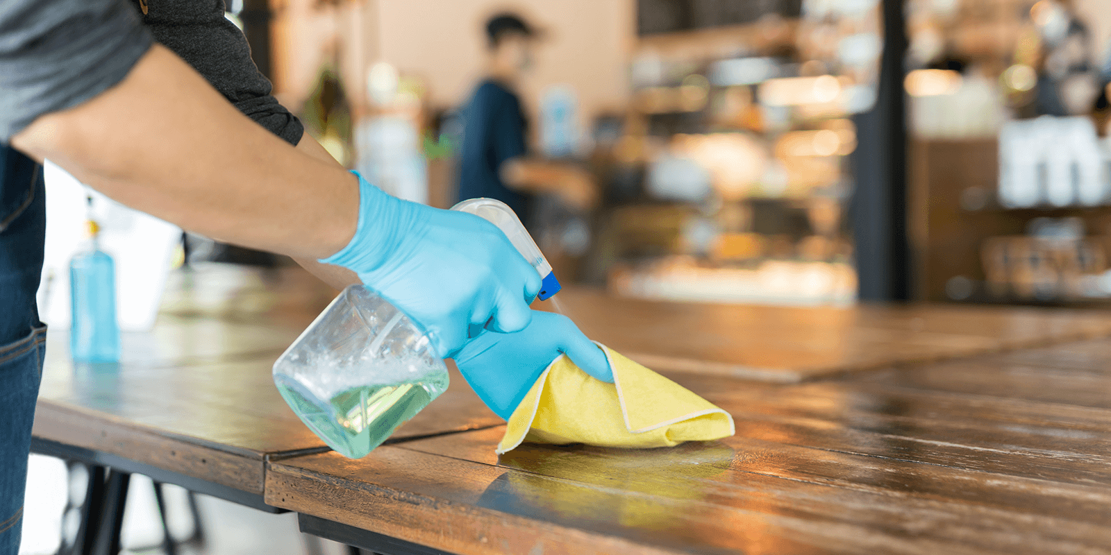 Person wearing blue gloves disinfecting a wooden table with spray and cloth in a cafe or restaurant.