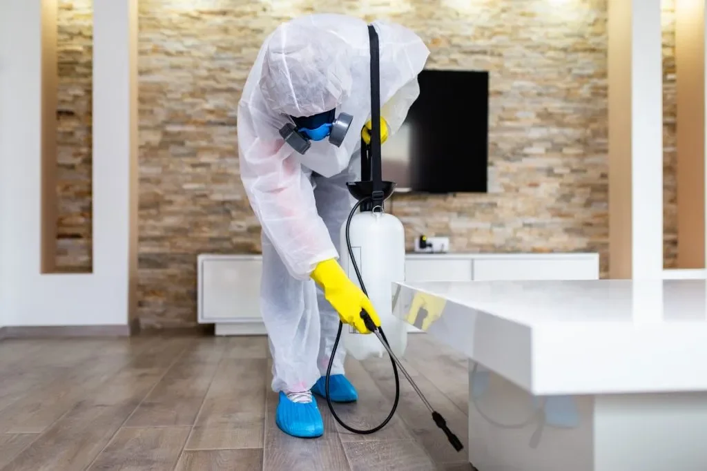Person dressed in protective suit using disinfectant spray in a modern indoor space.