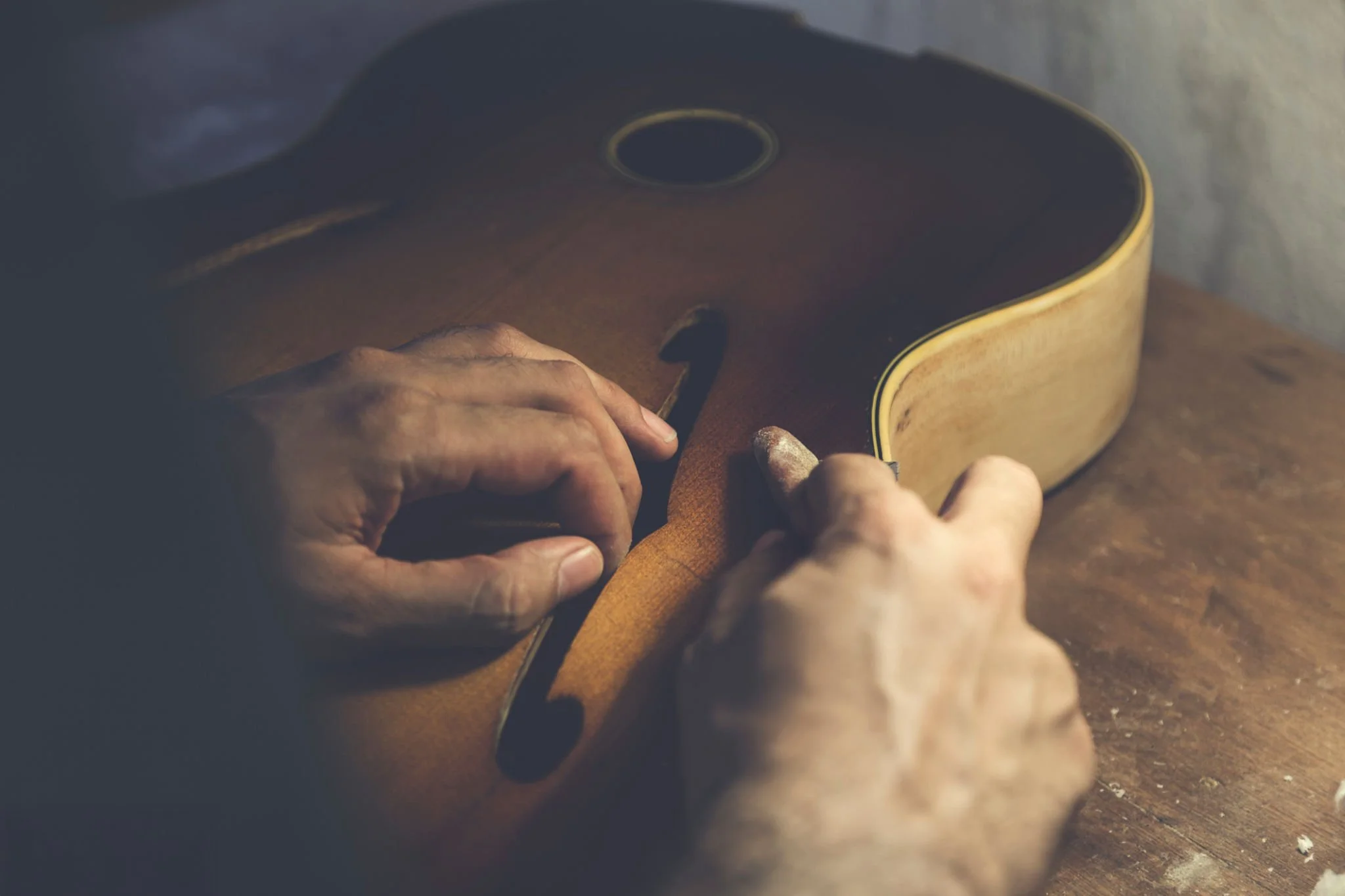 Close-up of hands crafting or repairing a wooden musical instrument, possibly an acoustic guitar, on a wooden surface.