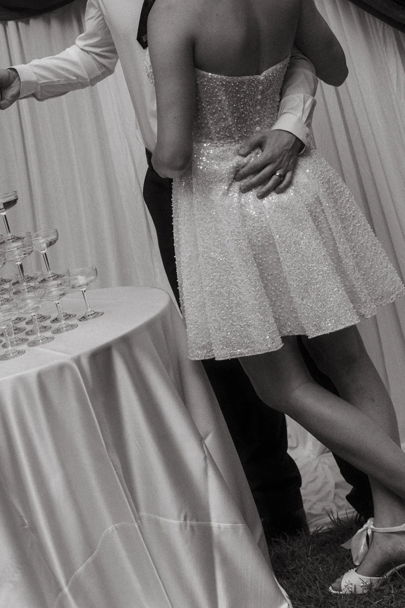 A black and white photo of a wedding dance, showing a bride in a sparkling dress and a groom in a suit, holding each other close, with a table of glasses in the foreground.