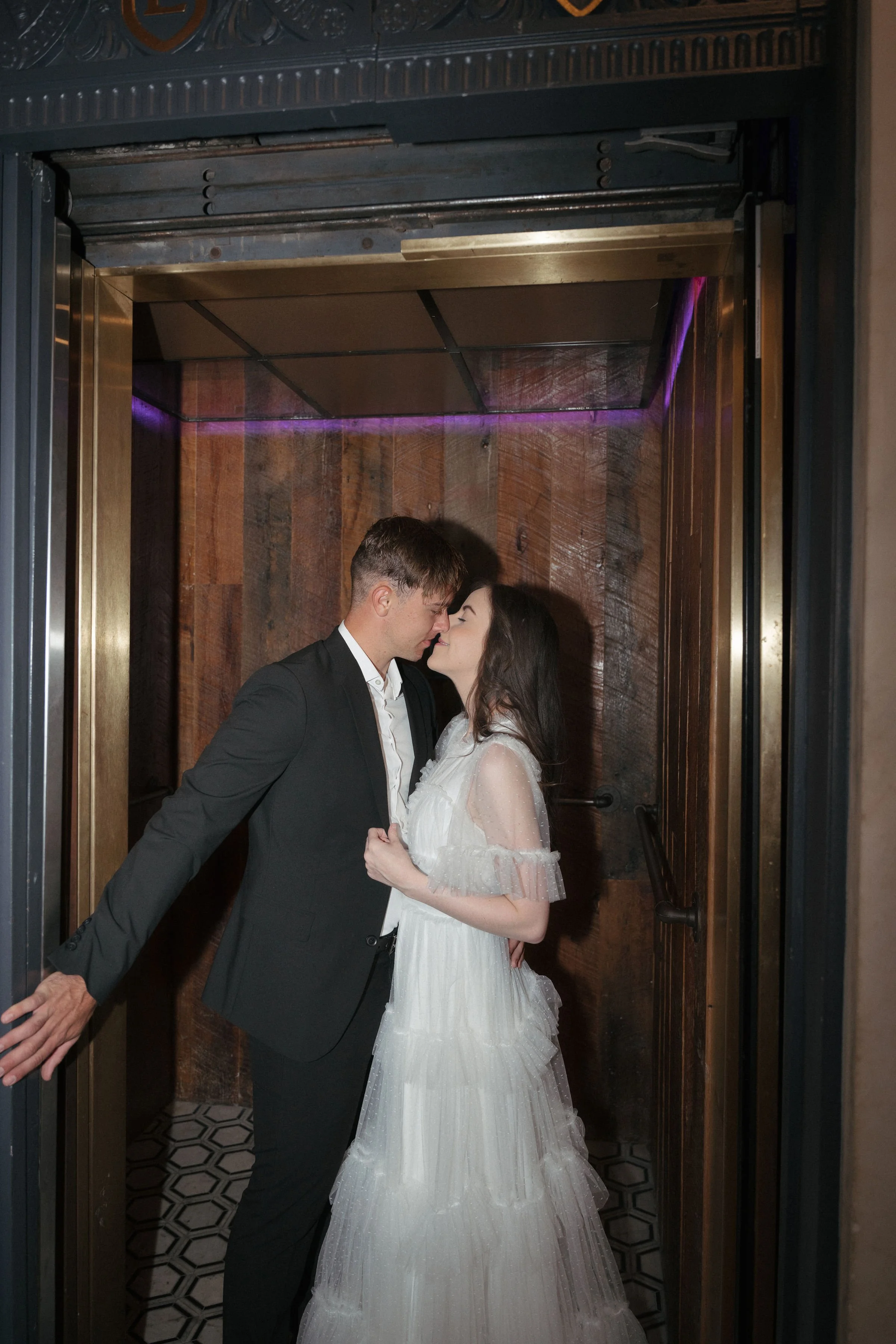 A couple in wedding attire sharing a kiss in an elevator with wooden paneling and purple lighting.