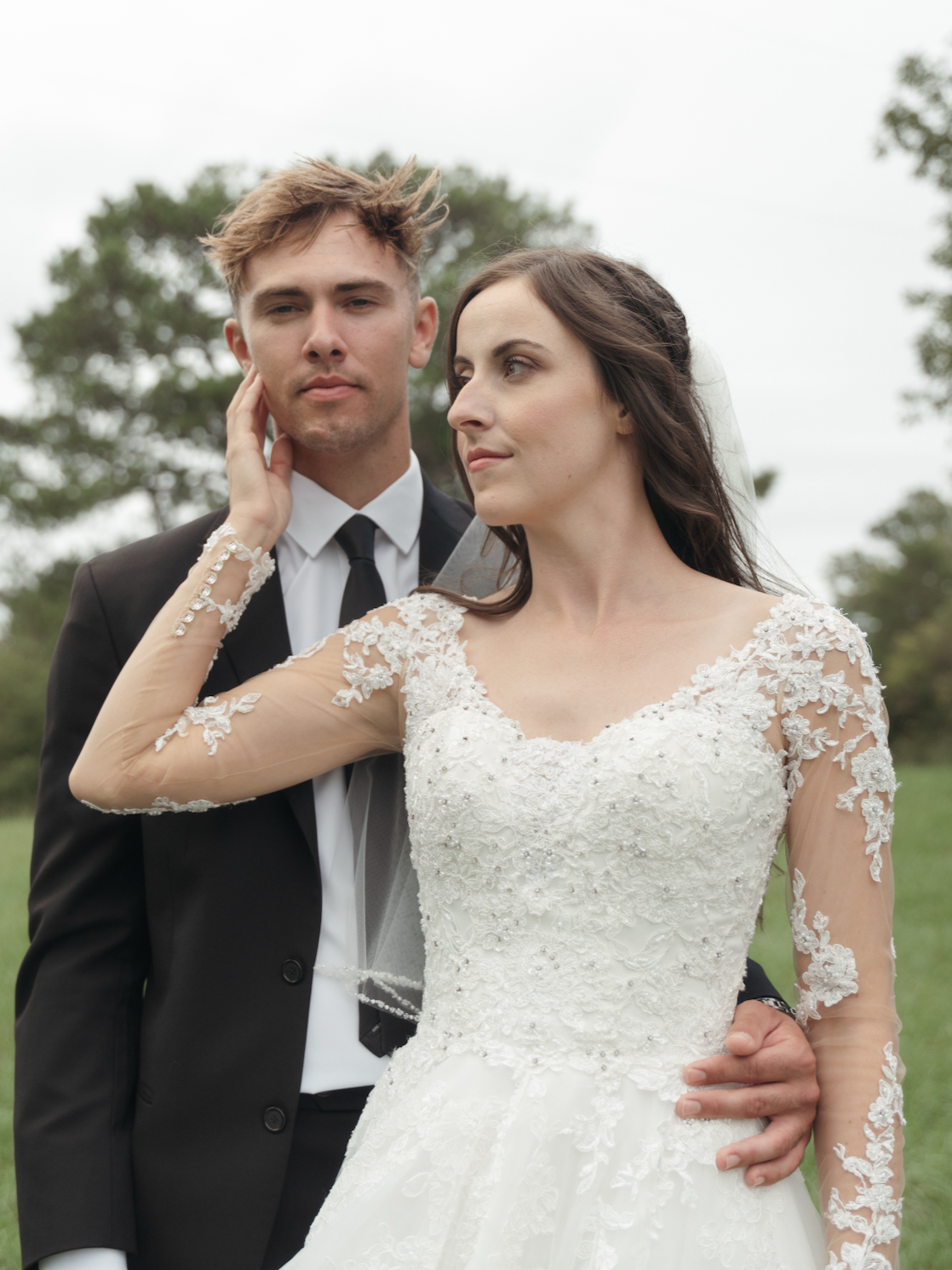 Bride and groom posing outdoors on wedding day, dressed in wedding gown and tuxedo, with trees in the background.