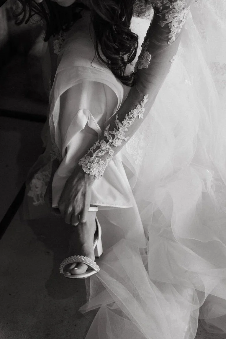 Close-up of a bride in a wedding dress, holding her high heel shoe while sitting.