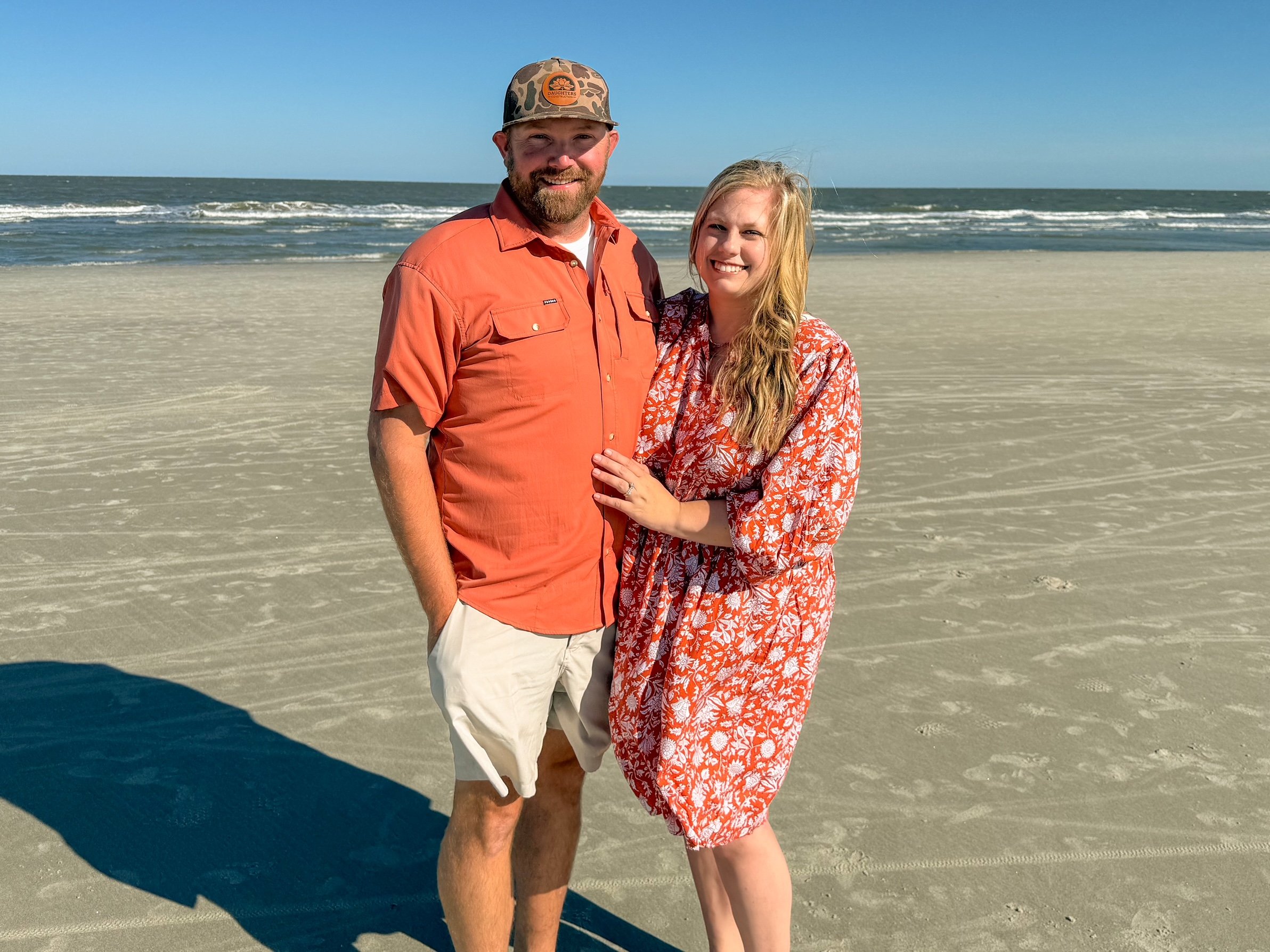 A man and a woman standing on a sandy beach with the ocean in the background. The man is wearing an orange shirt, beige shorts, and a camo hat, with a beard. The woman is wearing an orange floral dress, has long blonde hair, and is smiling. They are close together, with the woman’s hand resting on the man's arm.