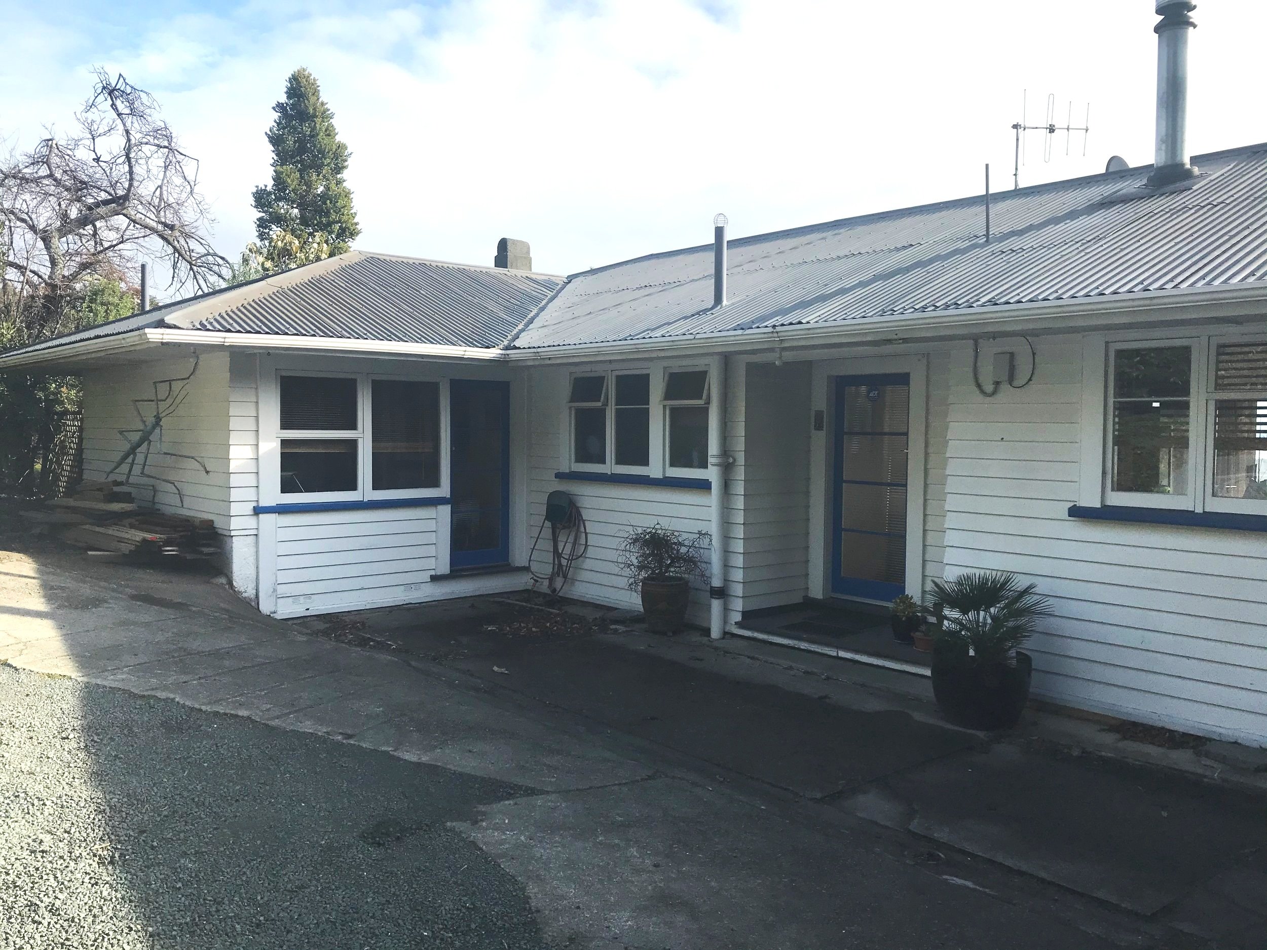 Front view of a white house with blue trims, multiple windows, a metal roof, and potted plants near the entrance, with a paved driveway and trees in the background.
