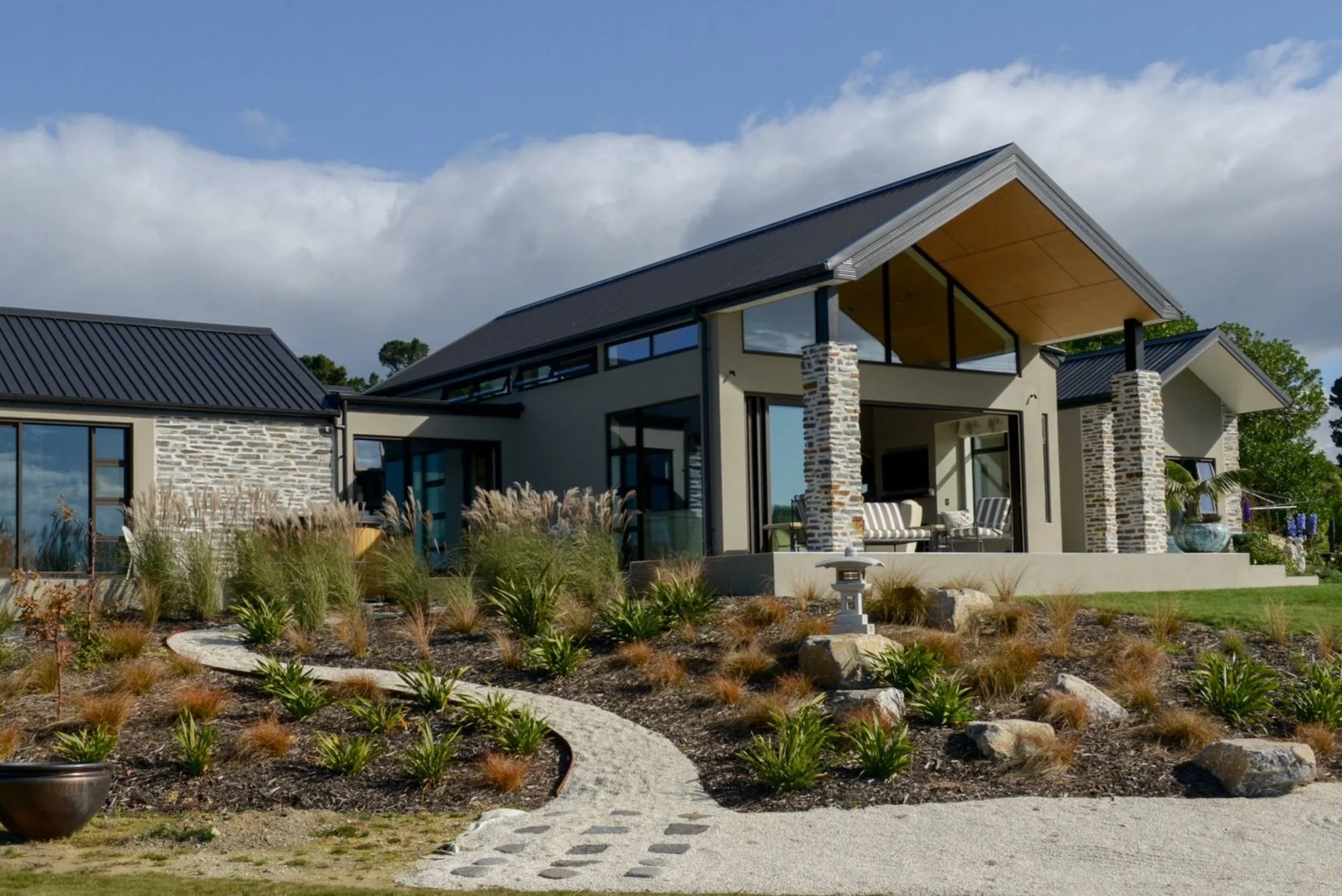 Modern architecturally designed house with a landscaped front yard, stone pillars, large glass windows, and a sloped roof, under a partly cloudy sky.