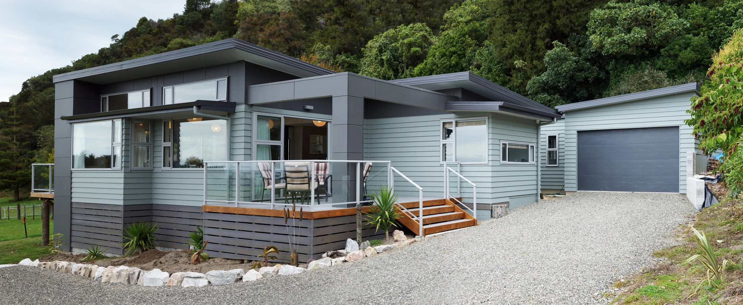 Modern house with gray exterior, attached garage, small deck with outdoor dining table, surrounded by landscaped yard and trees.