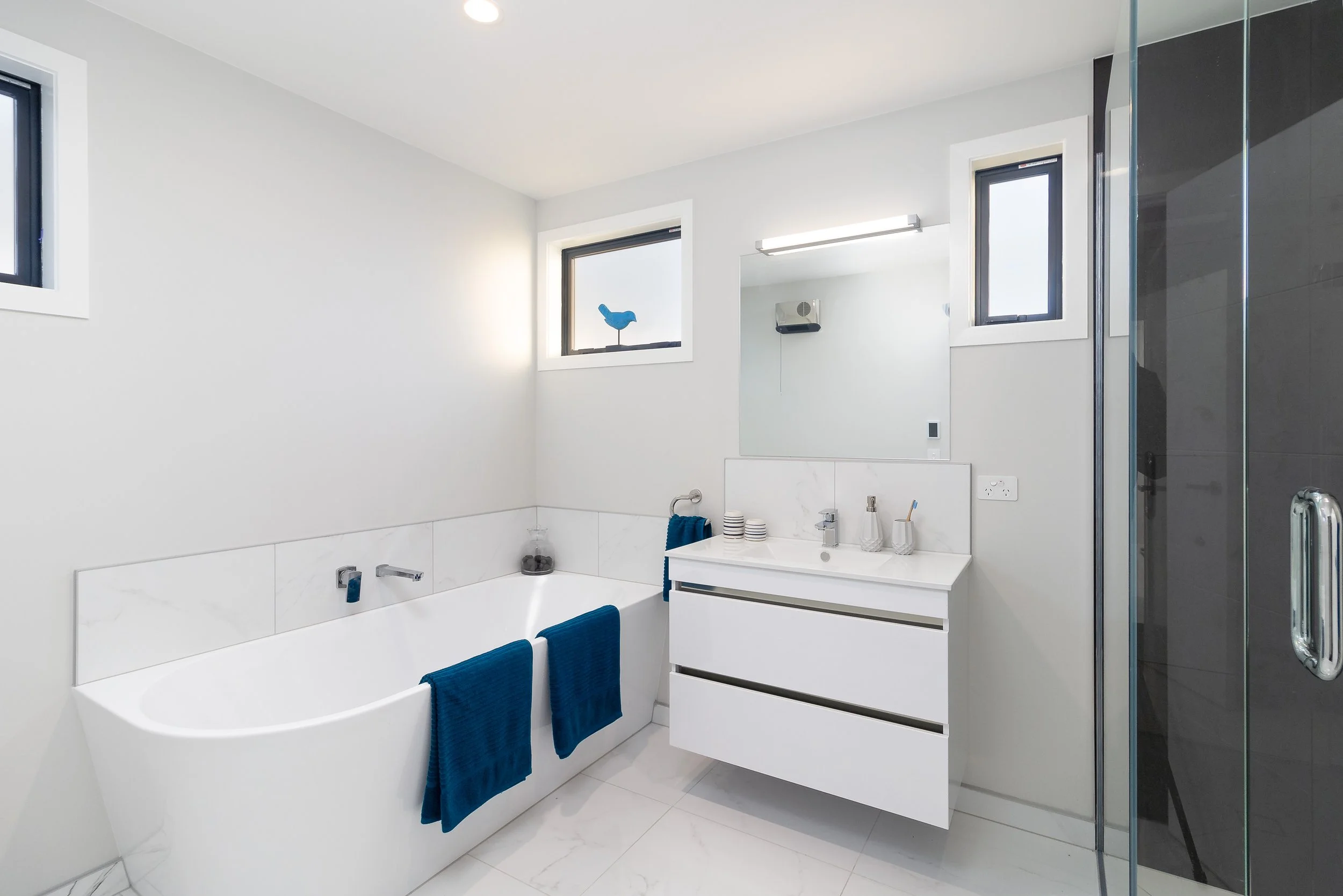 Modern architecturally designed white bathroom with a bathtub, vanity, mirror, and glass shower enclosure. Blue towels hang on the bathtub, and small windows allow natural light.