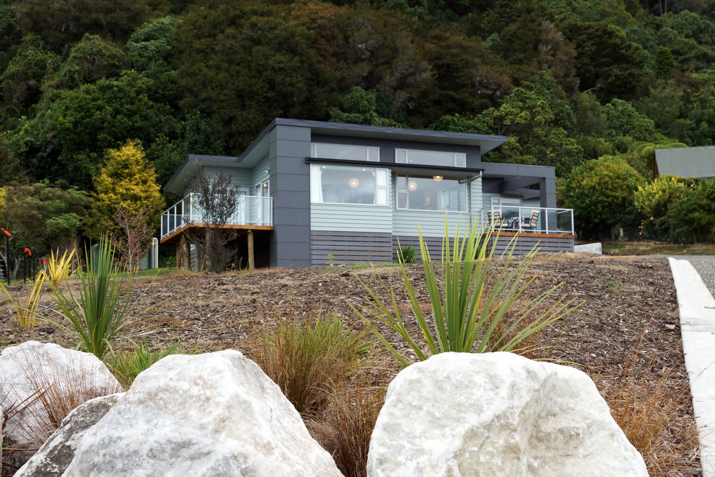 Modern house with large windows and balcony, situated on a hillside with autumn trees and rocks in the foreground.
