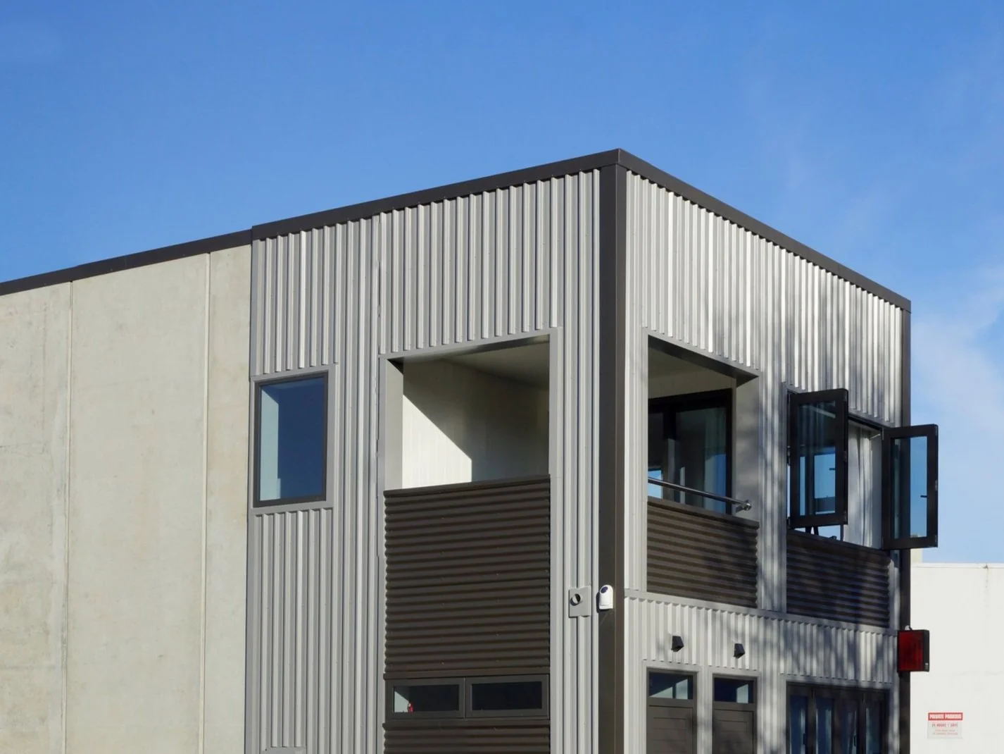 Modern multi-story building with vertical metal siding, rectangular windows, and open glass doors, against a clear blue sky.