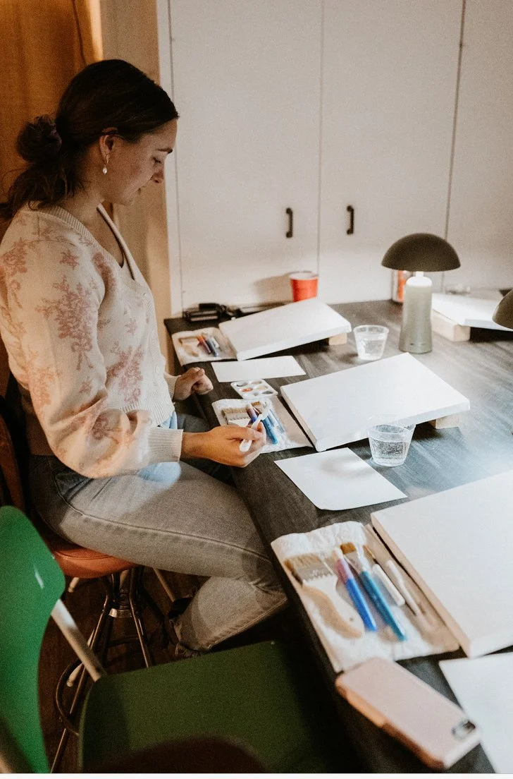 Woman sitting at a table, working with art supplies, including paint tubes, brushes, and sketchbooks, in a cozy room with white cabinets.
