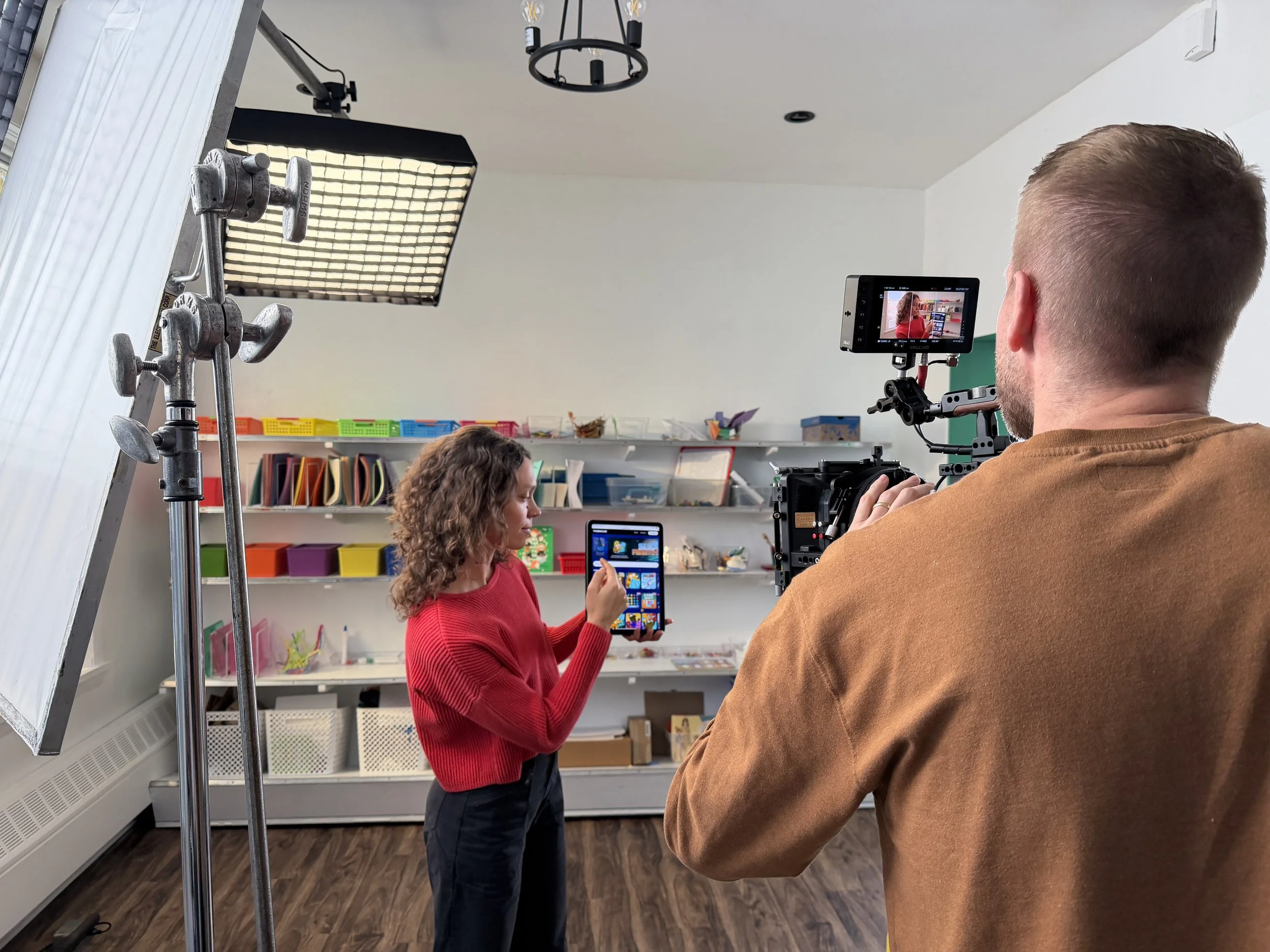 A woman in a red sweater is being filmed by a man in a brown shirt using professional filming equipment, including a camera and monitor, in a brightly lit classroom or studio with shelves of colorful storage bins and craft supplies in the background.