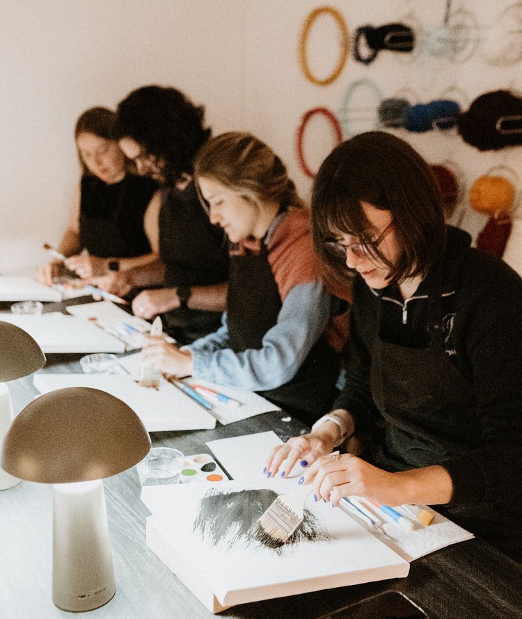 Group of five women painting and working on art projects at a table in a craft room, with wall hooks holding colorful yarn or string in the background.