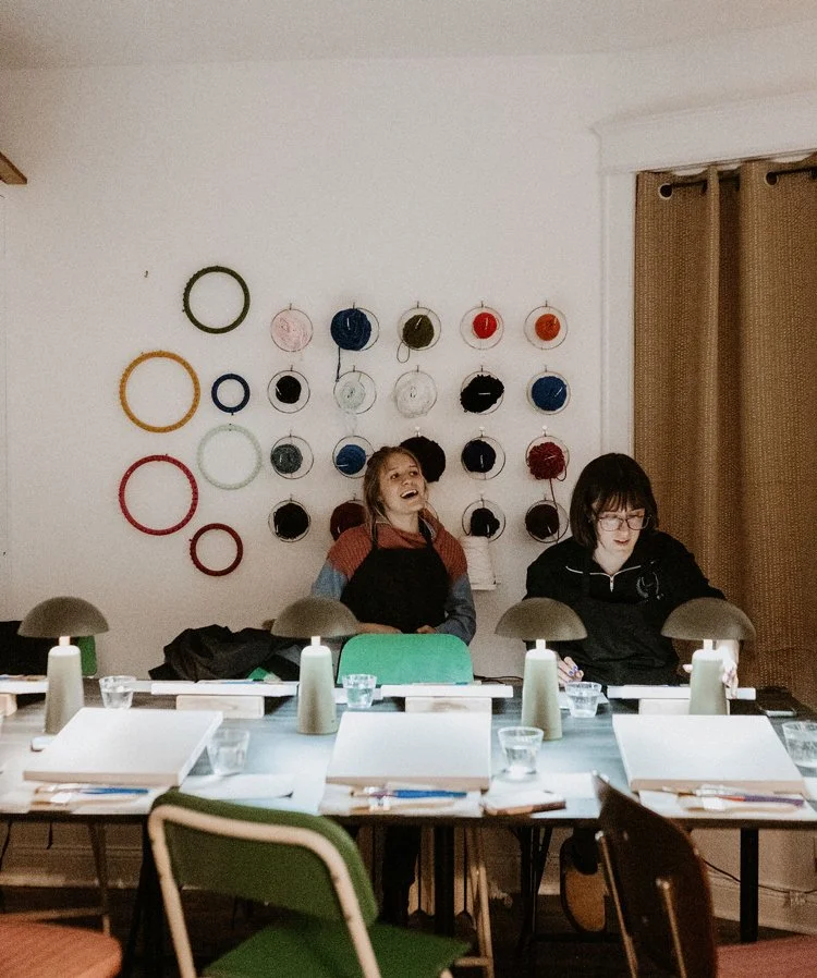 Two women sitting at a dinner table in a room with a wall decorated with yarn hoops in various colors and sizes.