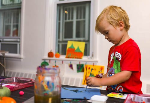 A young boy with blonde hair in a red shirt drawing with a marker at a table in a room decorated with fall-themed decorations and artwork.